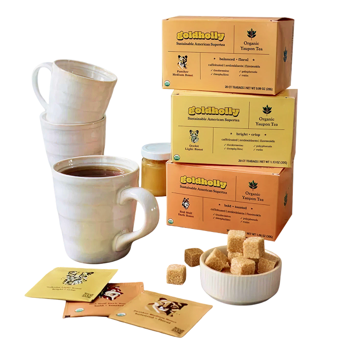 Goldholly organic yaupon tea three-roast collection — Light, Medium, and Dark Roast cartons stacked with white mugs, individual teabags, honey jar, and sugar cubes