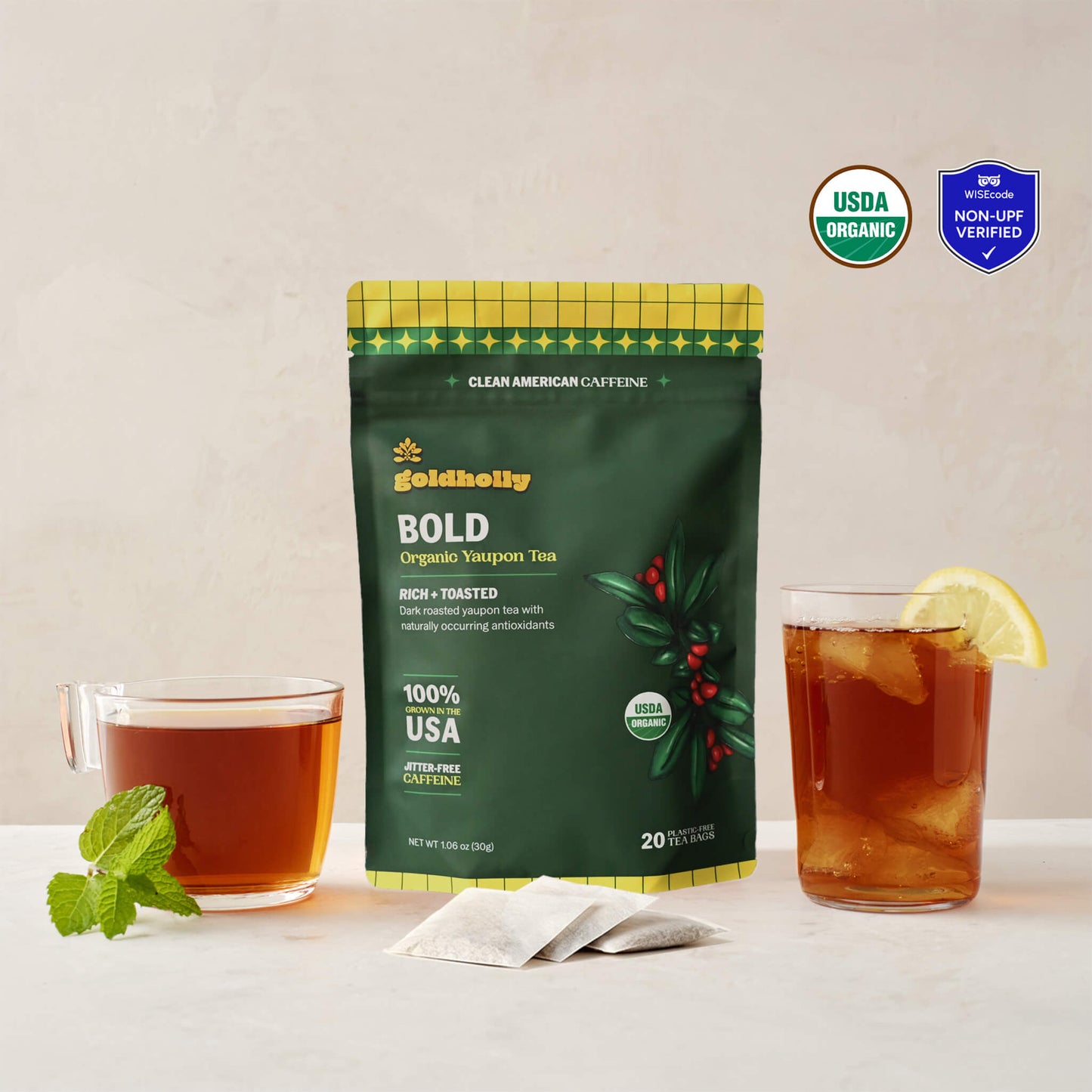 Goldholly Bold organic yaupon tea pouch in deep green with Rich + Toasted flavor tag, flanked by a warm cup of dark brewed tea with mint and an iced glass with lemon, with three teabags. USDA Organic certified and WISEcode Non-UPF verified.