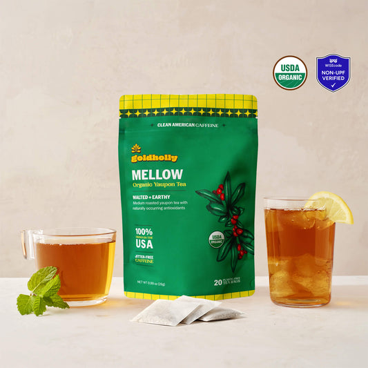 Goldholly Mellow organic yaupon tea pouch in vibrant green with Malted + Earthy flavor tag, flanked by a warm cup of brewed tea with mint and an iced glass with lemon, with three teabags. USDA Organic certified and WISEcode Non-UPF verified.