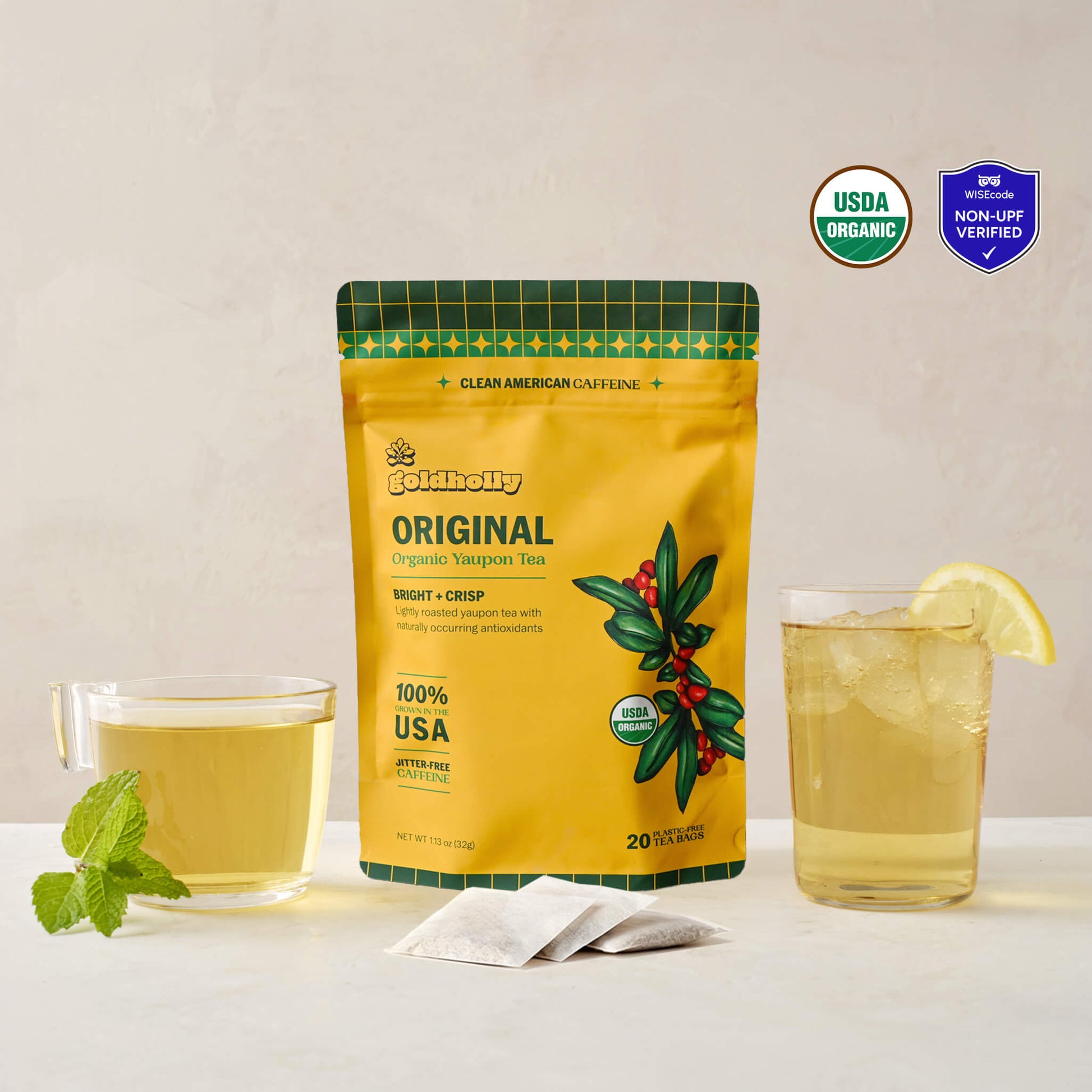 Goldholly Original organic yaupon tea pouch in golden yellow with Bright + Crisp flavor tag, flanked by a warm brewed cup with mint and an iced glass with lemon, with three teabags. USDA Organic certified and WISEcode Non-UPF verified.