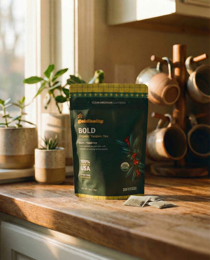 Goldholly Bold organic yaupon tea pouch on a warm sunlit kitchen counter with loose teabags beside it, copper mug tree and green plants in the background