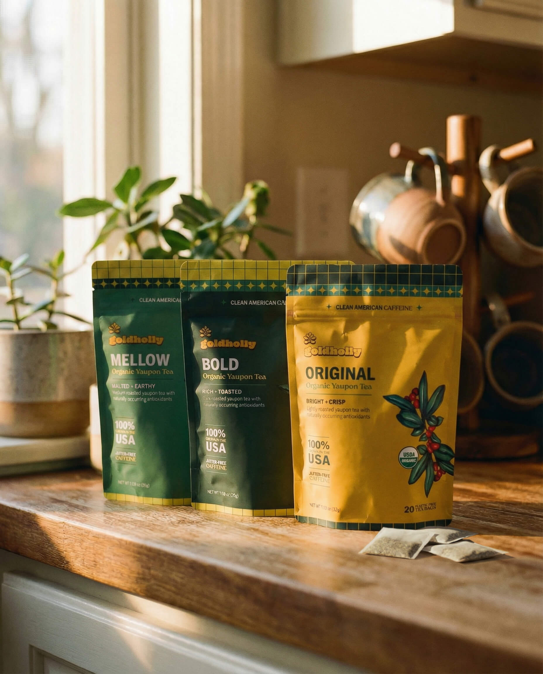Three Goldholly organic yaupon tea pouches, Original, Bold, and Mellow, lined up on a warm sunlit kitchen counter with loose teabags, copper mugs, and green plants