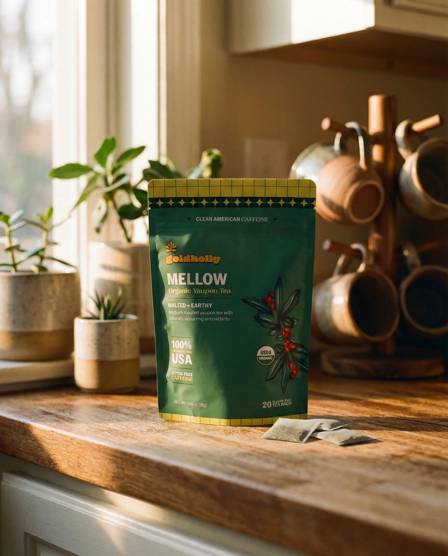 Goldholly Mellow organic yaupon tea pouch on a warm sunlit kitchen counter with loose teabags beside it, copper mug tree and green plants in the background