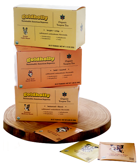 Goldholly organic yaupon tea three-roast collection stacked on a wood serving board — Light, Dark, and Medium Roast cartons with individual teabags displayed in front